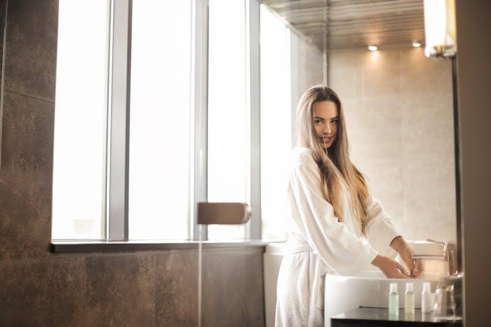 A woman standing near the tinted bathroom window, washing her hands at the sink.